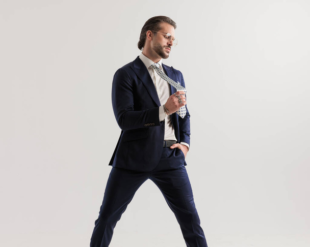 chladný elegantní podnikatel s brýlemi odvrací pohled při tahání kravatu a drží ruku v kapse na šedém pozadí - Fotografie, Obrázek
