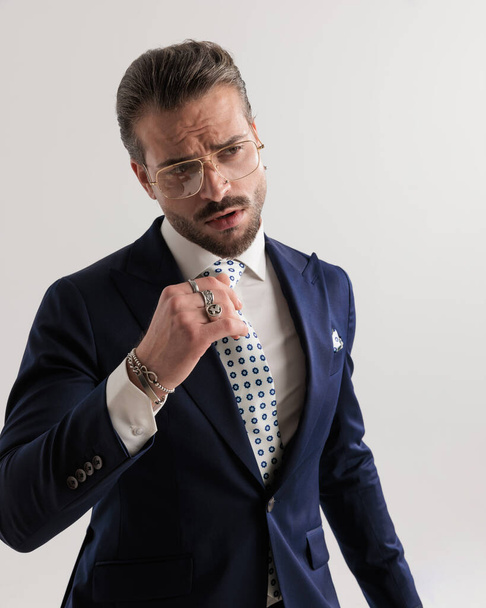 seksikäs korkealaatuinen liikemies laivastonsininen puku katselee pois, kun säädät solmiota harmaan taustan edessä  - Valokuva, kuva