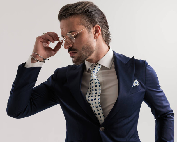 sexy barbudo hombre de negocios en elegante traje azul marino mirando hacia abajo lado y la organización de gafas en frente de fondo gris  - Foto, imagen