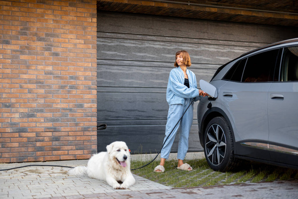 彼女の家のガレージの近くに彼女のかわいい犬と一緒に立っている間若い女性は彼女の電気自動車を充電します。現代的で幸せなライフスタイルの概念 - 写真・画像