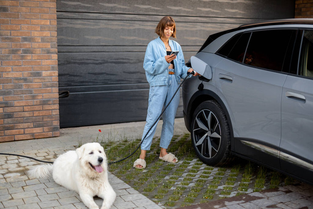 Νεαρή γυναίκα στέκεται με το σκύλο της και χρησιμοποιεί έξυπνο τηλέφωνο ενώ φορτίζει το ηλεκτρικό της αυτοκίνητο κοντά στο γκαράζ του σπιτιού της. Έννοια του σύγχρονου τρόπου ζωής και της βιωσιμότητας - Φωτογραφία, εικόνα