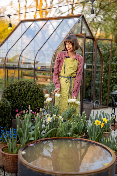 Porträt einer jungen Frau als Floristin, die in einem wunderschönen Garten mit Topfblumen, grünen Pflanzen und Vintage-Gewächshaus vor dem Hintergrund steht. Konzept Hobby und Gärtnern - Foto, Bild