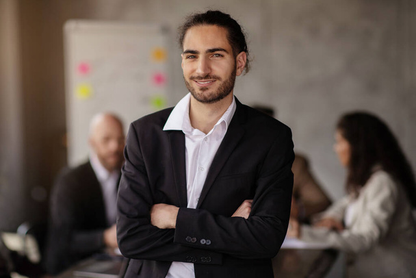 Предпринимательство. Успешный молодой бизнесмен уверенно позирует, стоя посреди современного офисного интерьера, улыбаясь в камеру и скрещивая руки. Бизнес карьера и лидерство - Фото, изображение