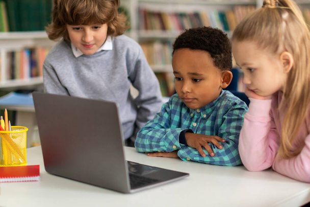 Διαφορετική ομάδα τριών ενθουσιωδών μαθητών που ασχολούνται με εκπαιδευτικές δραστηριότητες, εξερευνώντας προσεκτικά και μαθαίνοντας μαζί μέσω φορητού υπολογιστή στην τάξη του δημοτικού σχολείου - Φωτογραφία, εικόνα