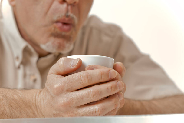 Amidst the cold embrace of a winter morning, the sun offers little relief. An aged man with an unrecognizable face tightly holds his coffee, blowing on it to cool. It's a delicate balance between warm - Photo, Image