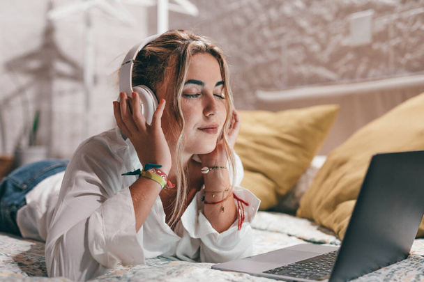 Közelkép egy fiatal, boldog nőről, aki laptopot használ otthon, és fülhallgatót visel, miközben zenét hallgat. Millenniumi tizenéves pihentető élvezi dalok csukott szemmel - Fotó, kép