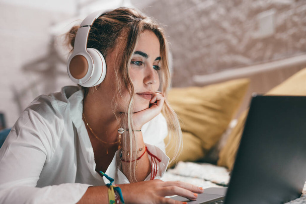 Close up of one young pretty happy woman using laptop indoor at home lying on the bed wearing headphones listening music. Millennial teenager having fun online studying. - Photo, Image