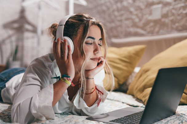 Κοντινό πλάνο μιας νεαρής αρκετά ευτυχισμένης γυναίκας που χρησιμοποιεί φορητό υπολογιστή στο σπίτι ξαπλωμένη στο κρεβάτι φορώντας ακουστικά ακούγοντας μουσική. Χιλιετής έφηβος χαλαρώνοντας απολαμβάνοντας τραγούδια με τα μάτια κλειστά - Φωτογραφία, εικόνα