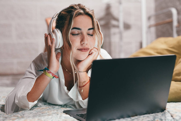 Lähikuva yksi nuori melko onnellinen nainen käyttää kannettavaa sisätiloissa kotona makaa sängyllä yllään kuulokkeet kuuntelee musiikkia. Millennial teini rentouttava nauttia kappaleita silmät kiinni - Valokuva, kuva