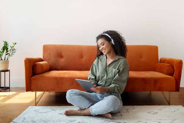 Rastgele tablet bilgisayar kullanan, kulaklık takan, internetten video izleyip hafta sonları modern oturma odasında dinlenen genç siyah bir kadın. İnternet, aygıt kavramı - Fotoğraf, Görsel
