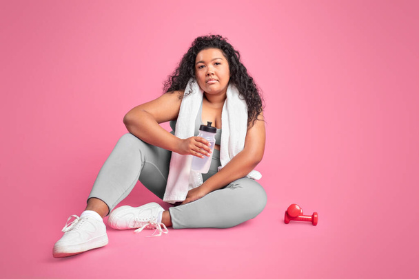 Усталое черное тело положительная женщина в спортивной одежде с полотенцем на шее, сидя на полу, держа бутылку воды, имеют перерыв после тренировки на розовом фоне студии, копировальное место - Фото, изображение