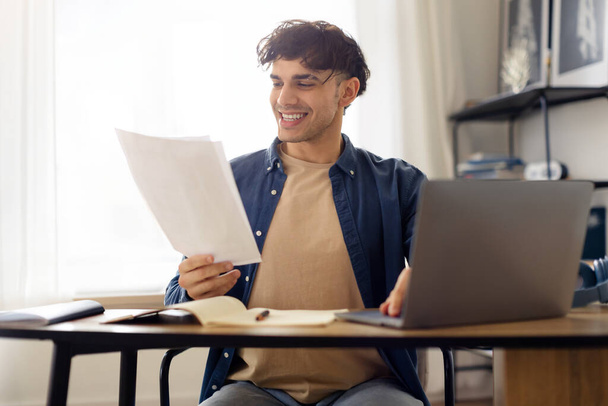 Papierkram. Fröhlicher junger Mann aus dem Nahen Osten, der Papierdokumente hält, Laptop benutzt und im Web surft, am Arbeitstisch im modernen Bürointerieur sitzt. Selektiver Fokus - Foto, Bild