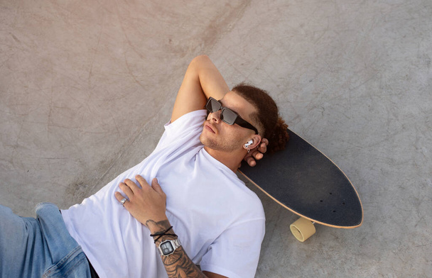 Jovem europeu encontra um momento de paz, deitado no seu skate, mergulhando na música através de auriculares ao ar livre, acima da vista - Foto, Imagem