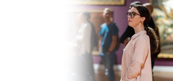 День музеев. Вид сбоку на молодую кавказскую женщину в очках и глядя на выставку. Люди и шедевры на заднем плане. Веб-баннер с макетом. - Фото, изображение
