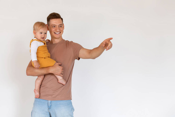 Spójrz tam. Radosny młody mężczyzna trzymający niemowlę i wskazujący obok stojąc razem na tle białej ściany, Millennial Father wiążący się ze swoim dzieckiem w domu, Copy Space - Zdjęcie, obraz