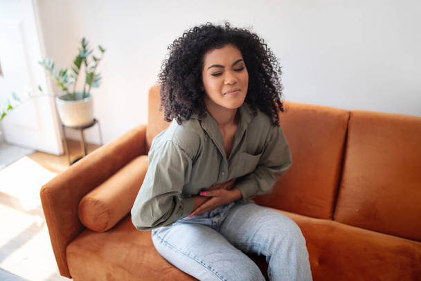 Gastritis Disease. Black woman suffering from stomachache, touching her stomach while having painful spasm in belly sitting on couch at home. Abdominal pain symptom, health problems concept - Photo, Image
