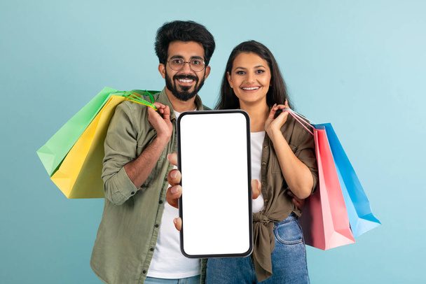 Веселые красивые молодые индийские пара женщина и мужчина держит покупки, показывая большой смартфон с белым экраном и улыбаясь, используя хороший торговый мобильное приложение, синий фон, копировальное пространство - Фото, изображение