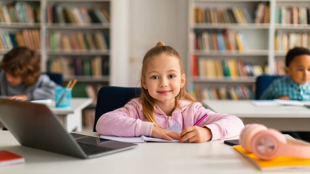 Vrolijk Europees schoolmeisje zit aan haar bureau, bezig met laptop en schrijven in copybook, glimlachen naar de camera, beeltenis van een vrolijke en positieve educatieve ervaring, panorama - Foto, afbeelding