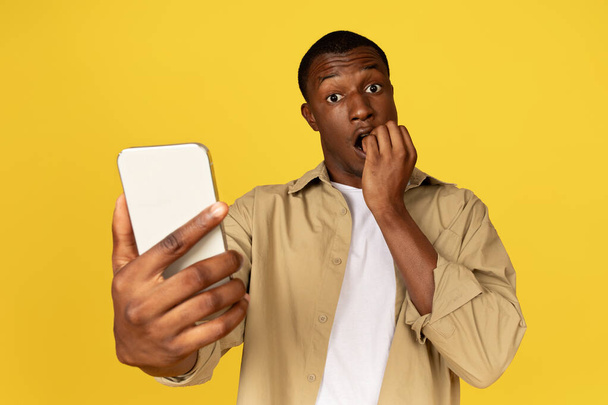 Shocked scared millennial african american guy in casual bite fingers, look at smartphone, has bad news, isolated on yellow studio background. Ad and offer, human emotions, stress reaction - Photo, Image