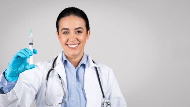 Professional latin woman doctor in protective gloves, confidently exhibits syringe to camera, preparing for injection against grey studio background, panorama, free space - Photo, Image