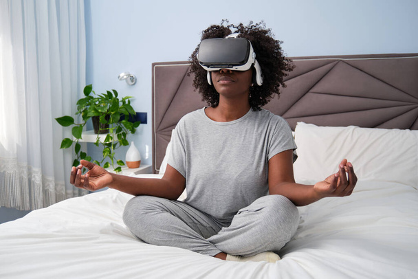 Mujer joven africana con auriculares de realidad virtual meditando, disfrutando de la simulación y relajándose en el dormitorio. - Foto, Imagen