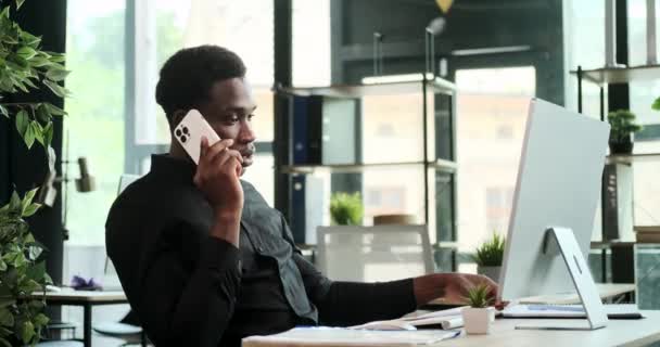 El gerente afroamericano participa en una conversación telefónica mientras está en la estación de trabajo. Profesionalismo y enfoque en la comunicación efectiva son evidentes en esta escena de eficiencia en el lugar de trabajo. - Imágenes, Vídeo