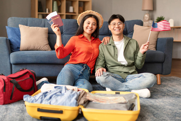 Весела корейська пара тримає прапор США та упаковку валізи вдома, посміхаючись на камеру, показуючи свої паспорти та квитки. Молоді подружжя готуються подорожувати або можливо переїзд - Фото, зображення
