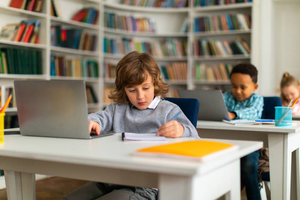 Akıllı okul çocukları, ilköğretim sınıfındaki masalarda oturuyorlar, dizüstü bilgisayarlarla eğitim içeriğini araştırıyorlar, dijital öğrenme dünyasını araştırıyorlar, eğitime çağdaş bir yaklaşımı işaret ediyorlar. - Fotoğraf, Görsel