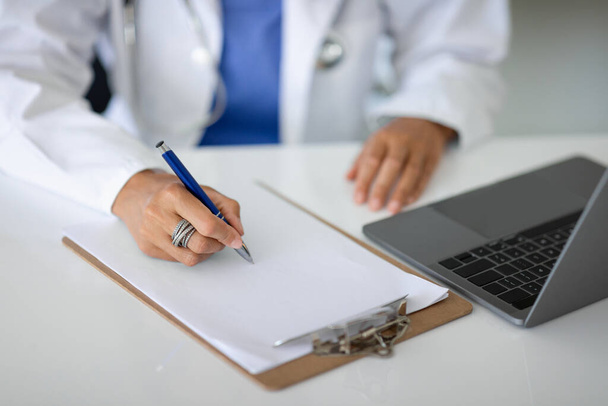 Обрезанный женщина в медицинской форме врач сидит за столом с ноутбуком компьютер, делает заметки в медицинской карте во время консультации с пациентом. Концепция здравоохранения - Фото, изображение