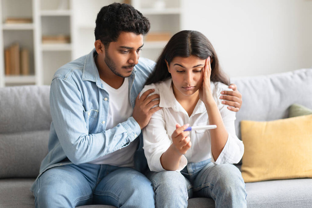 Ungewollte Schwangerschaft. Besorgtes Hindu-Paar mit Blick auf positives Testergebnis, gestresste indische Männer und Frauen, die nicht bereit sind, Eltern zu sein, zu Hause auf dem Sofa sitzend - Foto, Bild