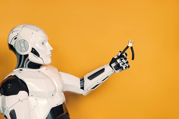 Un robot sosteniendo alicates en su mano está aislado sobre un fondo naranja. Concepto de maestro robot. Colaboración entre humanos e inteligencia artificial. - Foto, imagen