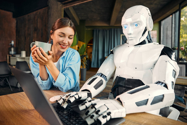 Νεαρή γυναίκα με ένα φλιτζάνι καφέ και ένα ανθρωποειδές ρομπότ που εργάζεται ενώ κάθεται σε ένα φορητό υπολογιστή σε ένα σύγχρονο γραφείο. Συνεργασία μεταξύ ανθρώπων και τεχνητής νοημοσύνης. - Φωτογραφία, εικόνα