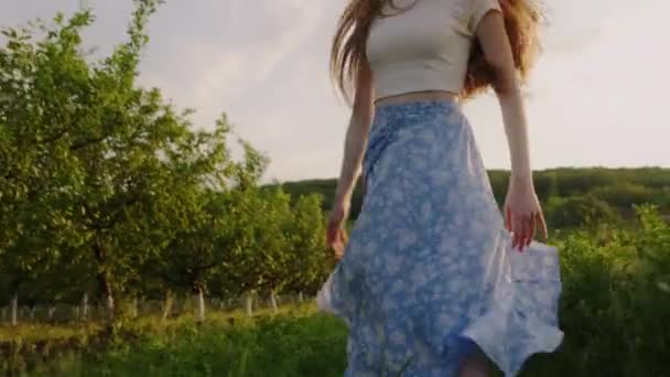 バックビューファッショナブルなケアフリー美女ランニングブルースカートを着て,花の田舎の庭で楽しんでいます. 元気な若い女の子が歩いている - 映像、動画