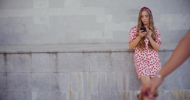 Blondynka młoda kobieta za pomocą smartfona opierając się na ścianie zatrzymującej z mężczyzną chodzącym z przodu - Materiał filmowy, wideo