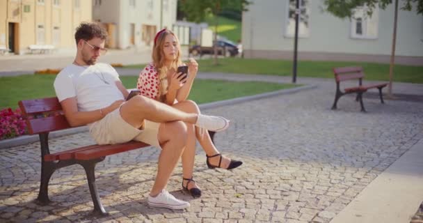 Heterosexuelles Paar sendet SMS auf Mobiltelefonen, während es im Park auf einer Bank sitzt - Filmmaterial, Video