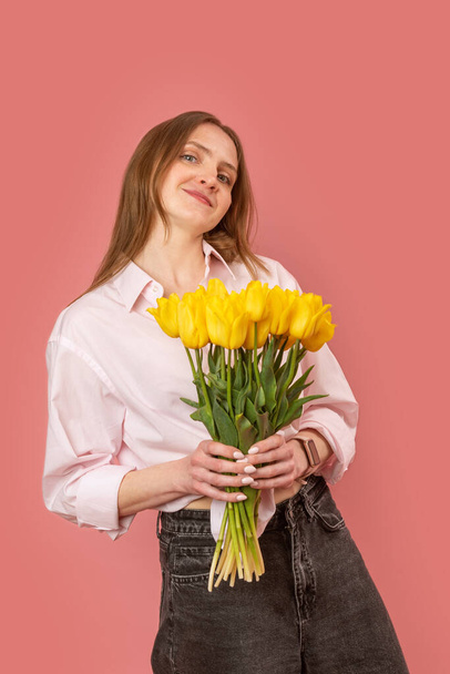 Young beautiful European woman spring bouquet of yellow flowers tulips in her hands. Isolated on pink background. women day. Mother day. Vertical frame - Photo, Image