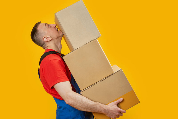 Мужчина средних лет в рабочей форме держит большие тяжелые коробки. Портрет погрузчика в студии на желтом фоне. Вид сбоку. Копирование пространства, макет - Фото, изображение