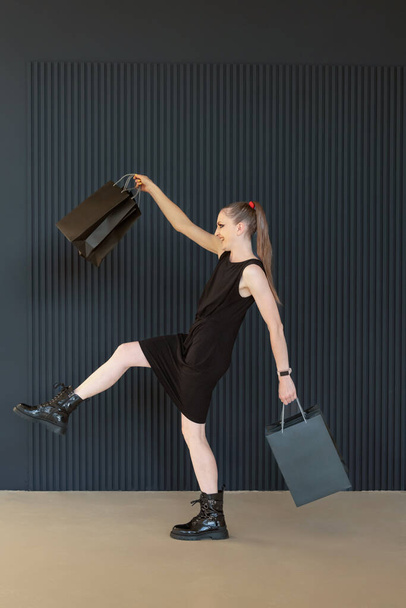 Fiatal nő fekete ruhában fekete bevásárló táskák vesz egy hosszú lépés, szezonális kedvezmény. Teljes hosszúságú portré, függőleges keret. Fekete péntek, eladások. - Fotó, kép