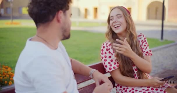 Szőke fiatal nő nevet, miközben beszél férfi barátja ül padon a parkban - Felvétel, videó