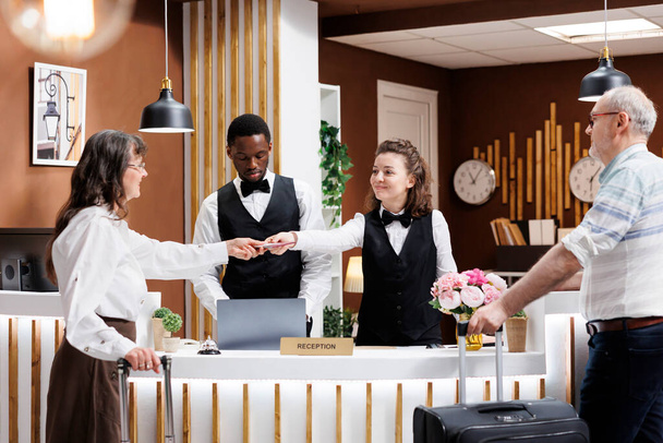 Freundlicher Concierge hilft älteren männlichen und weiblichen Touristen in der Hotellobby. Rentnerin stellt notwendige Dokumente für den Check-in bereit, während Reisender am Registrierungsschalter geduldig wartet. - Foto, Bild
