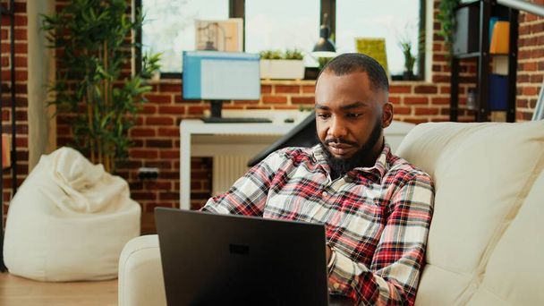 ソファに横たわっている間、リラックスしたアフリカ系アメリカ人のテレワーカーはノートパソコンで彼の電子メールをチェックします。明るいスタイリッシュな居心地の良いアパートで自宅から働くリモート従業員個人事務所 - 写真・画像