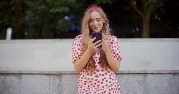 Mujer rubia riendo mientras mensajes de texto en el teléfono inteligente contra la pared de contención - Metraje, vídeo