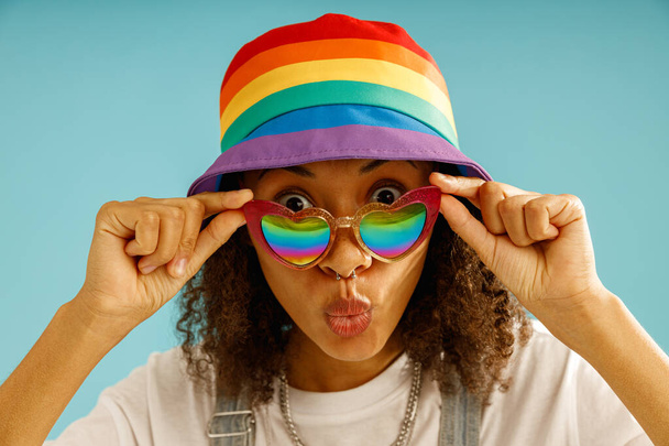 虹の帽子とハート型のメガネの笑顔の女性は青いスタジオの背景にカメラを見ます - 写真・画像