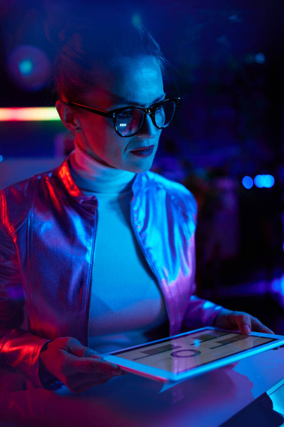 Neon metaverse futuristic concept. pensive modern business woman in glasses using tablet PC and exploring charts in modern office. - Foto, afbeelding