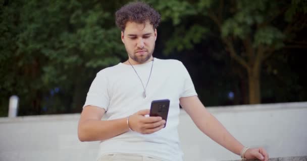 Usmívající se mladý muž textové zprávy na mobilním telefonu, zatímco stojí v parku - Záběry, video