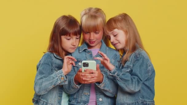 Teenage dívky používají mobilní smartphone psaní procházení říct Wow ano zjistil velkou výhru dobrou zprávu oslavit, hraní online hry. Sestřičky. Tři sourozenci děti na žlutém pozadí - Záběry, video