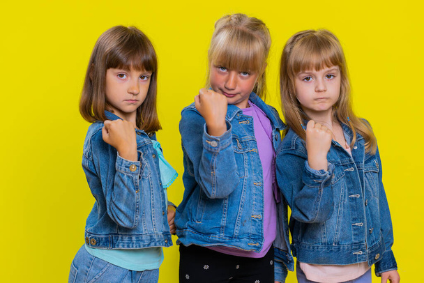 Aggressive angry teenage girls trying to fight at camera, shaking fist, boxing with expression, punishment, abuse, bullying. Little children sisters. Three siblings kids isolated on yellow background - Photo, Image