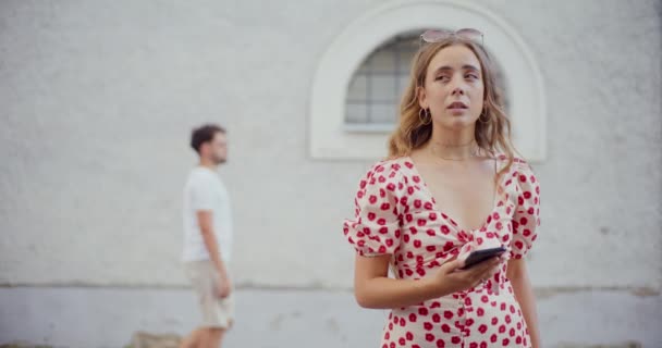 Bedachtzame blonde vrouw met smart phone met man lopen in de achtergrond tegen gebouw - Video