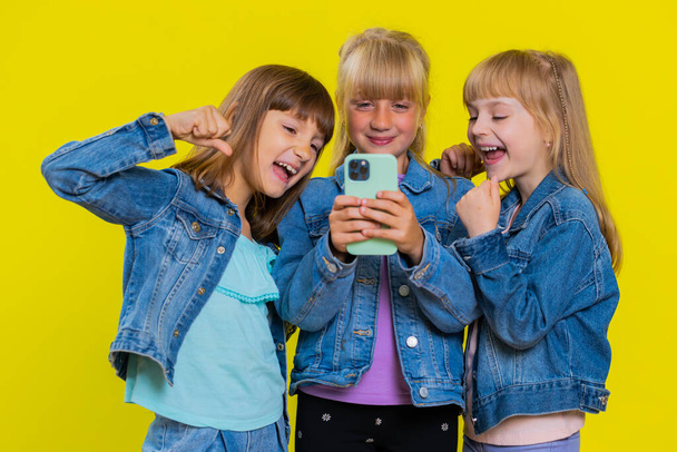 Teenage girls use mobile smartphone typing browsing say Wow yes found out great big win good news celebrate, playing online game. Little children sisters. Three siblings kids on yellow background - Photo, Image