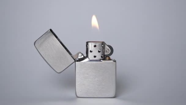 Un encendedor de metal gris se quema a la luz sobre un fondo gris. La llama del encendedor parpadea lentamente. Reflejos de llama en el encendedor de hierro. - Metraje, vídeo